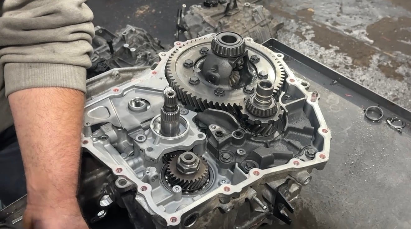 Mechanic holding disassembled automotive transmission showing internal gears and components on workbench